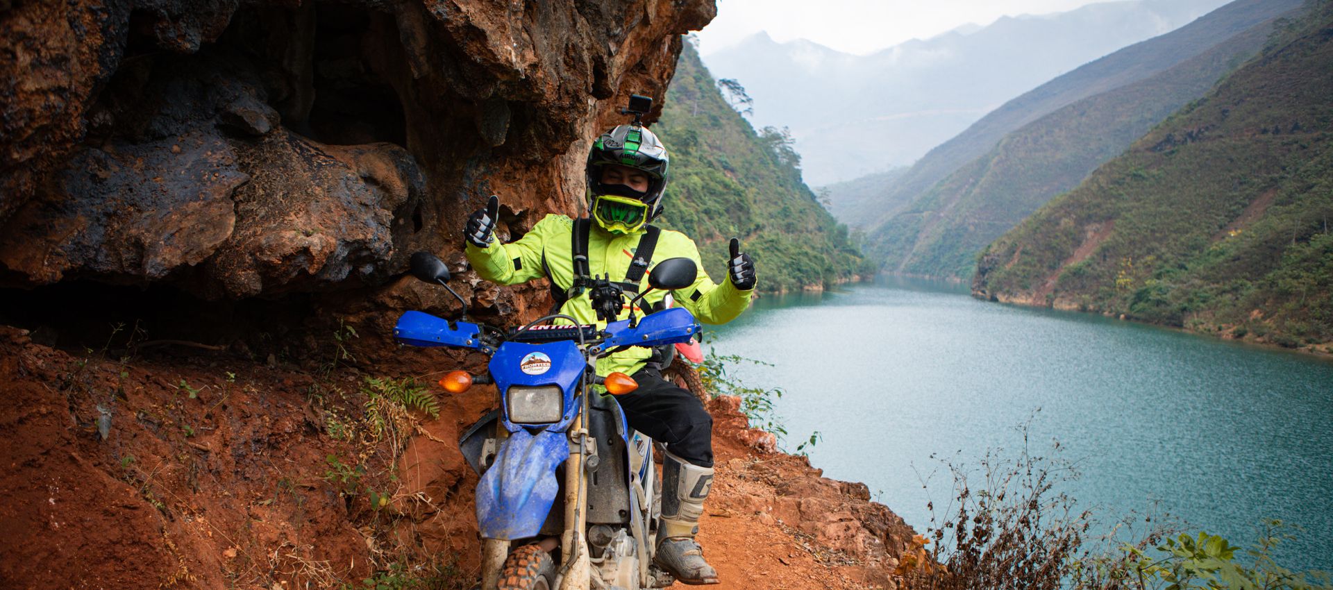 Motorcyclist in bright gear giving thumbs up while on a narrow cliff path with lake view