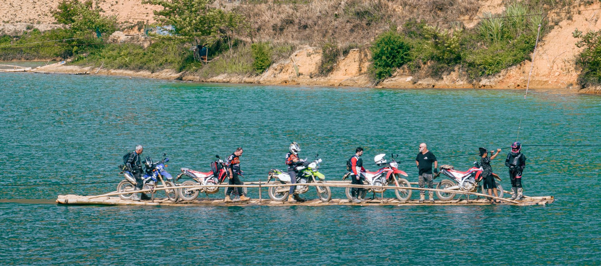 Group of motorcyclists with their bikes crossing a river on a wooden raft