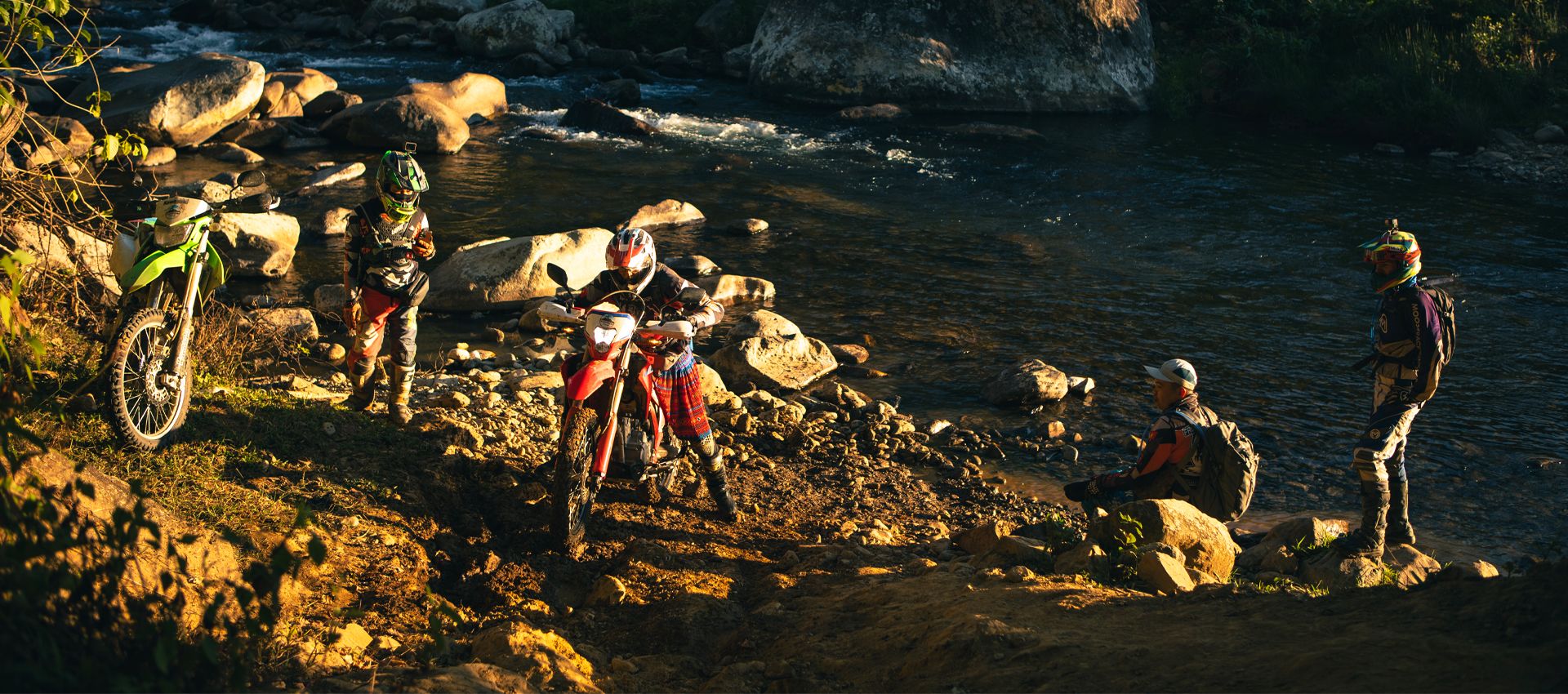 Motorcyclists relaxing by a river with dirt bikes in golden hour lighting